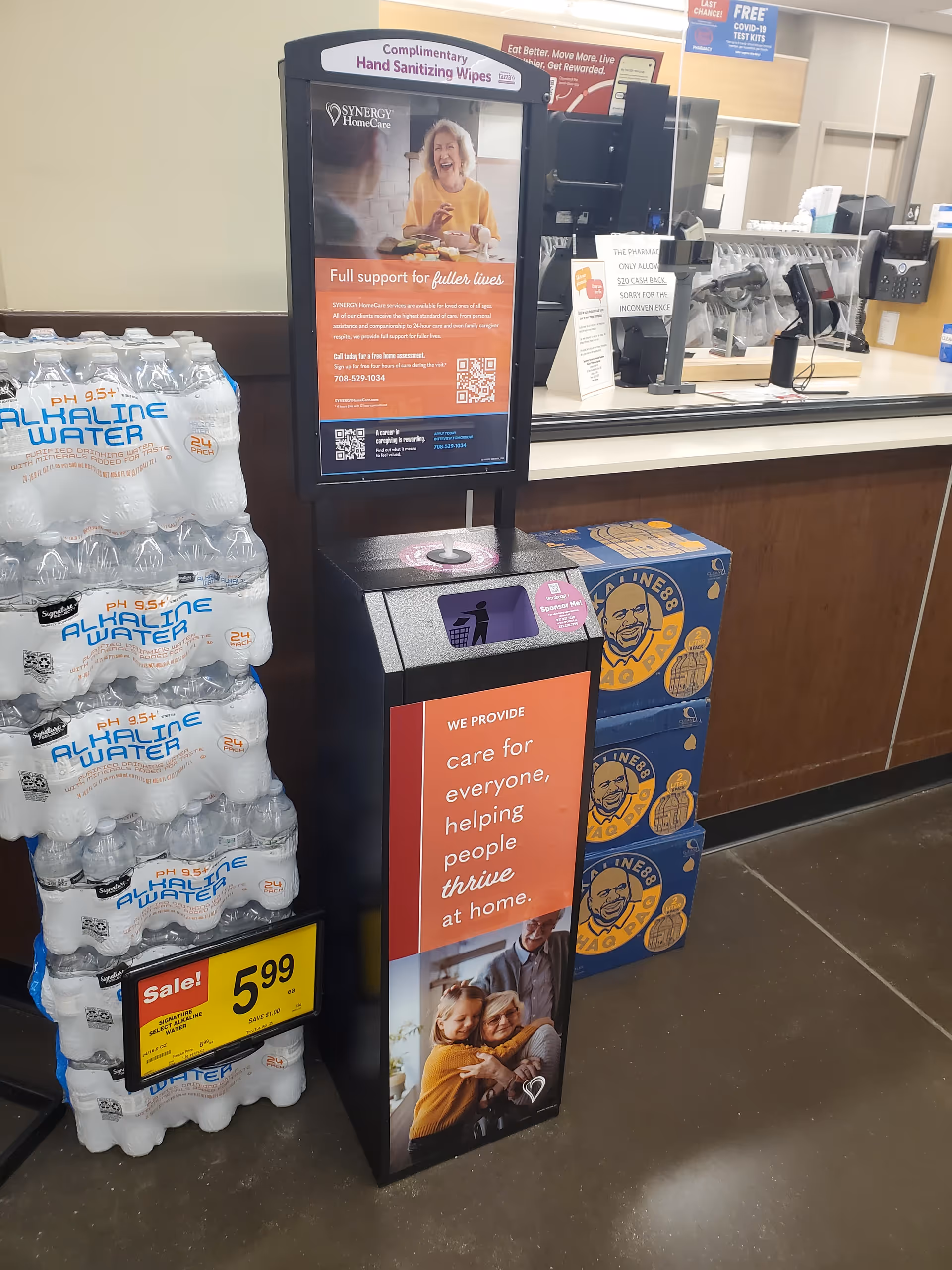 Reception area with a SYNERGY HomeCare poster beside a counter and bottled water stacks