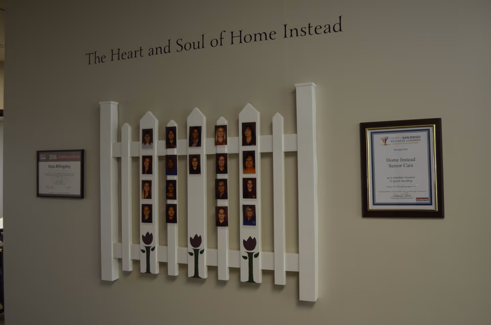Interior wall display featuring portrait photos and certificates beneath The Heart and Soul of Home Instead.