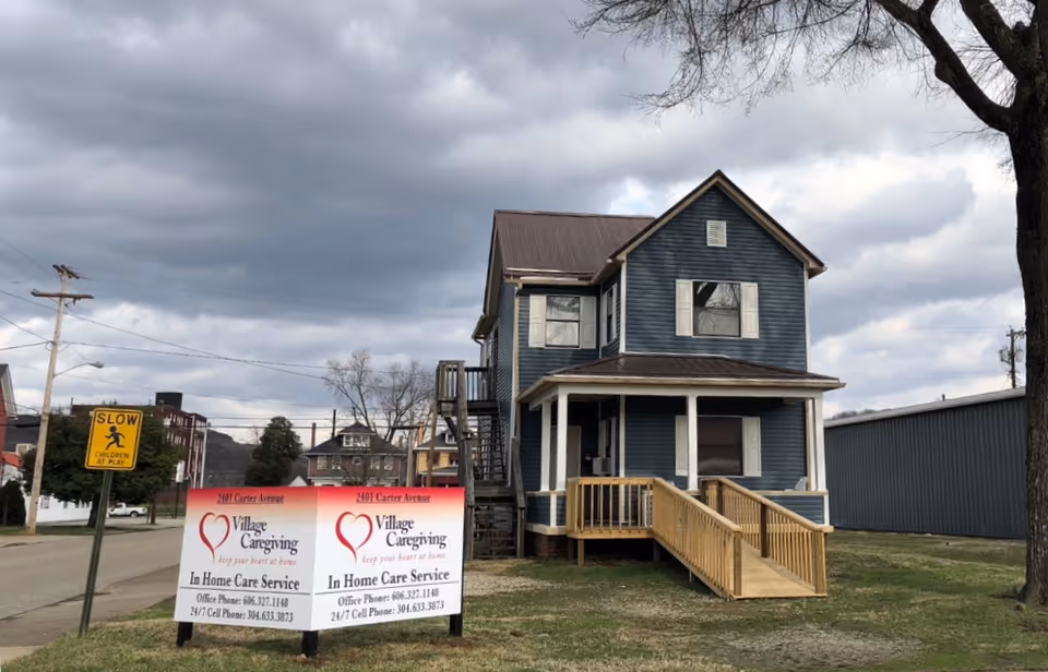 Blue two-story house-style building with a wooden ramp and Village Caregiving sign in yard