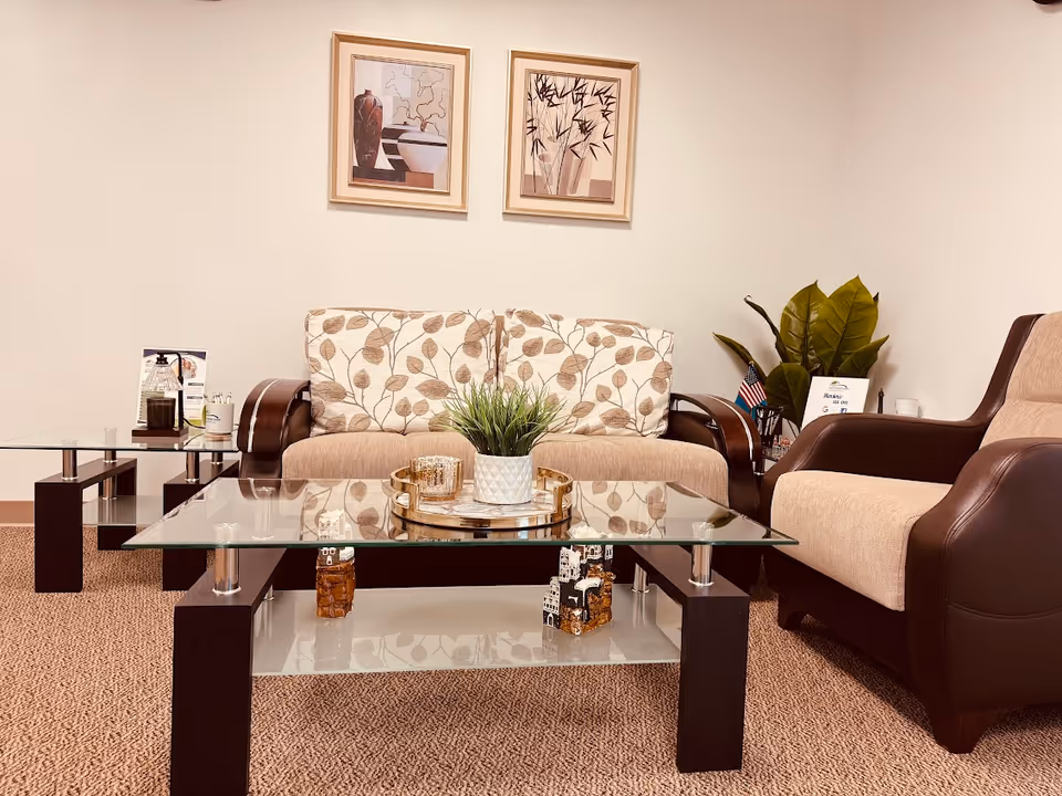 A cozy senior living facility lounge with glass coffee tables, cushioned sofa set, plants, and art.