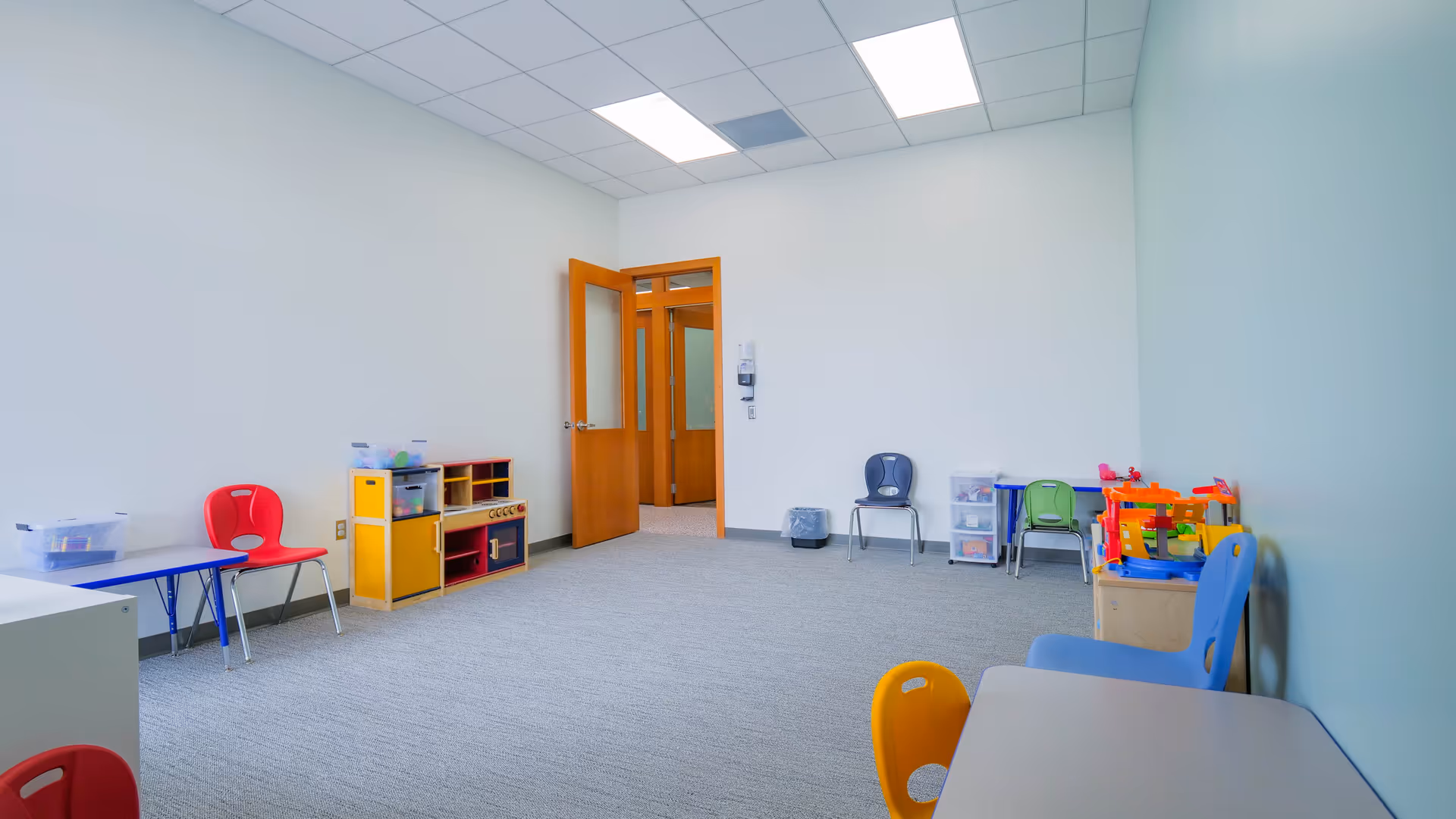 An empty children's playroom with small tables, chairs, and toy shelves along pale walls.