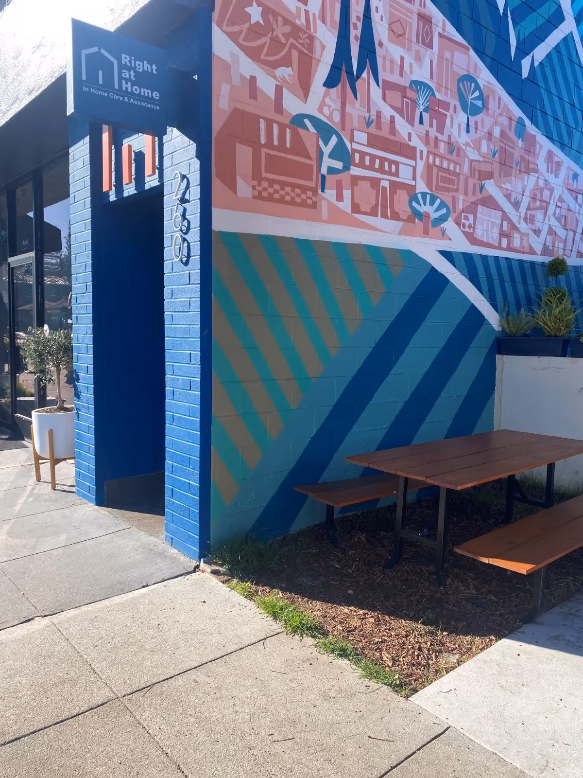 Exterior view of Right at Home facility with blue brick wall, mural, and wooden picnic table.