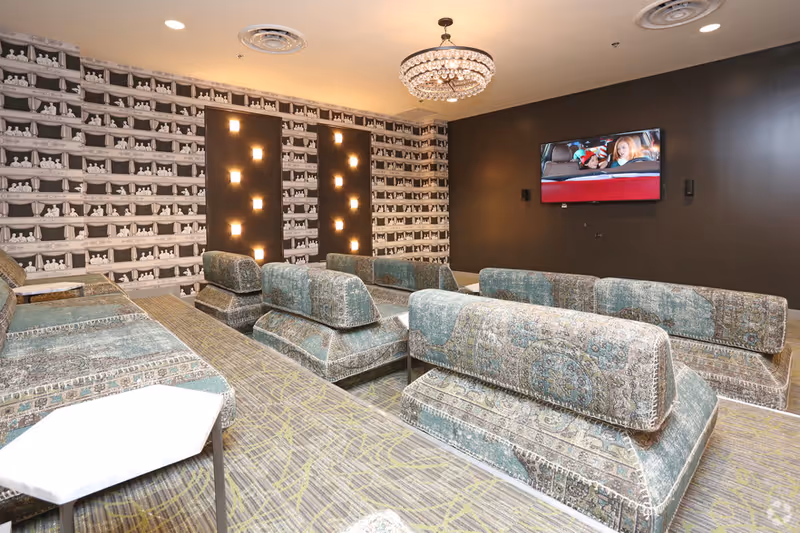 A spacious residents' lounge with tiered blue patterned couches, a chandelier, and a wall-mounted TV.