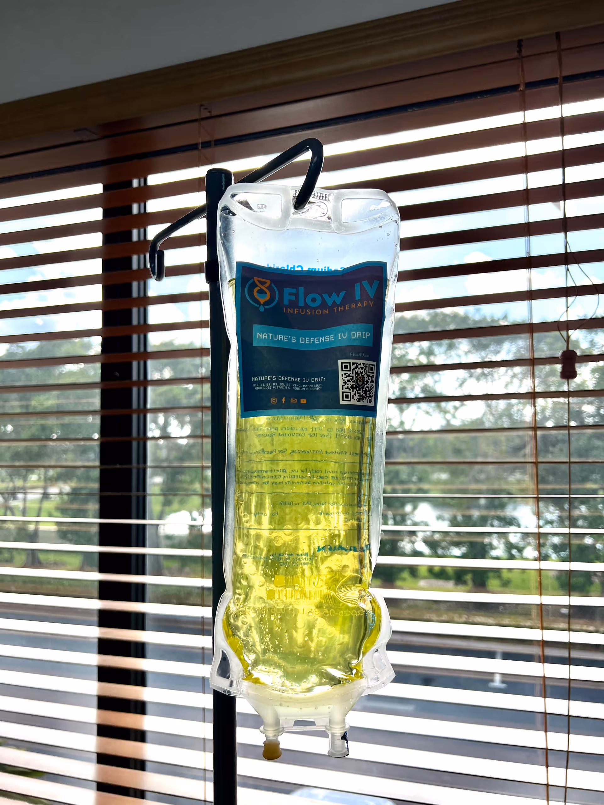 A Flow IV infusion bag hangs on a stand indoors near window blinds.