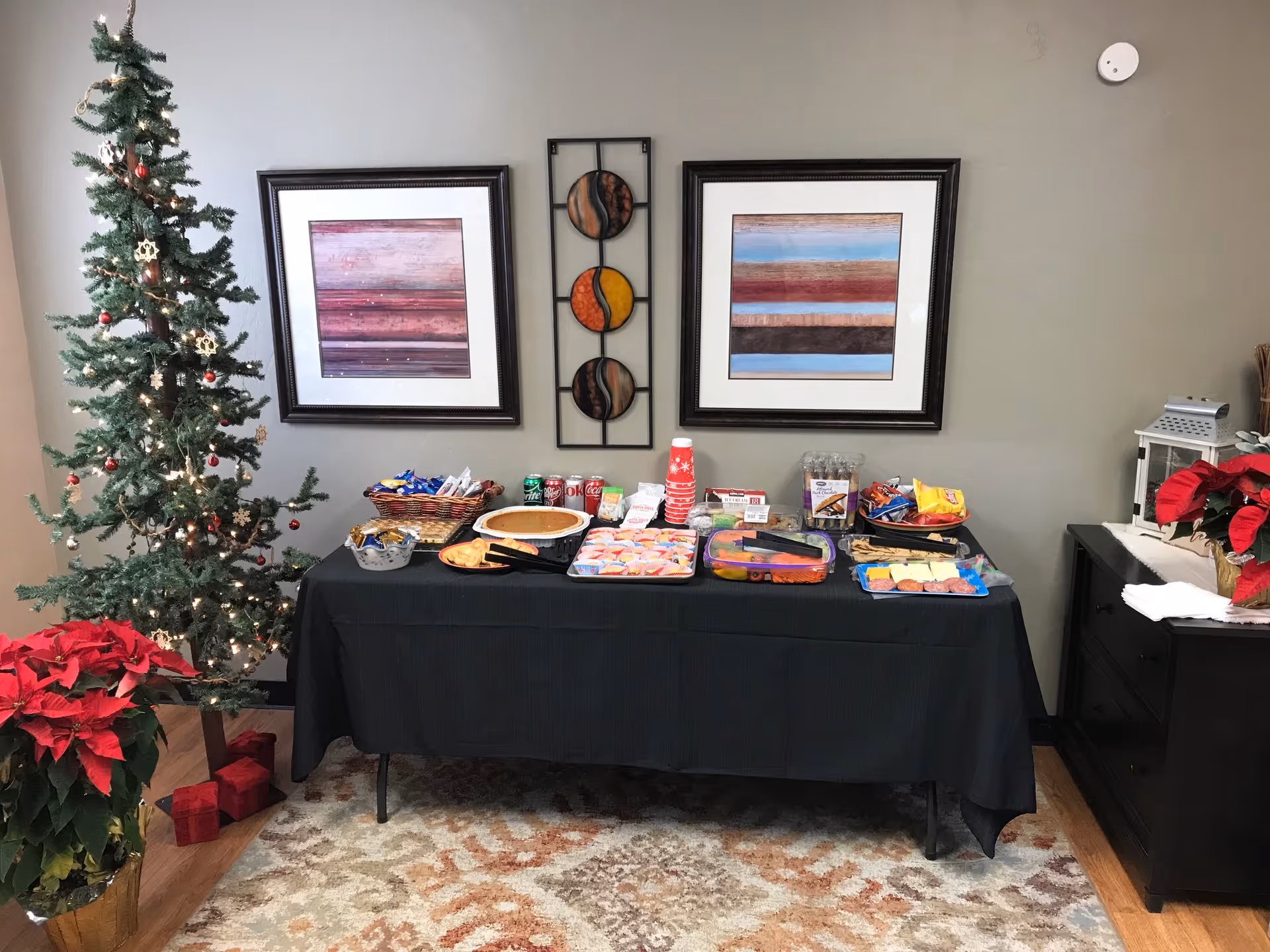 A snack buffet table with beverages, holiday decor, and framed wall art inside a living area.