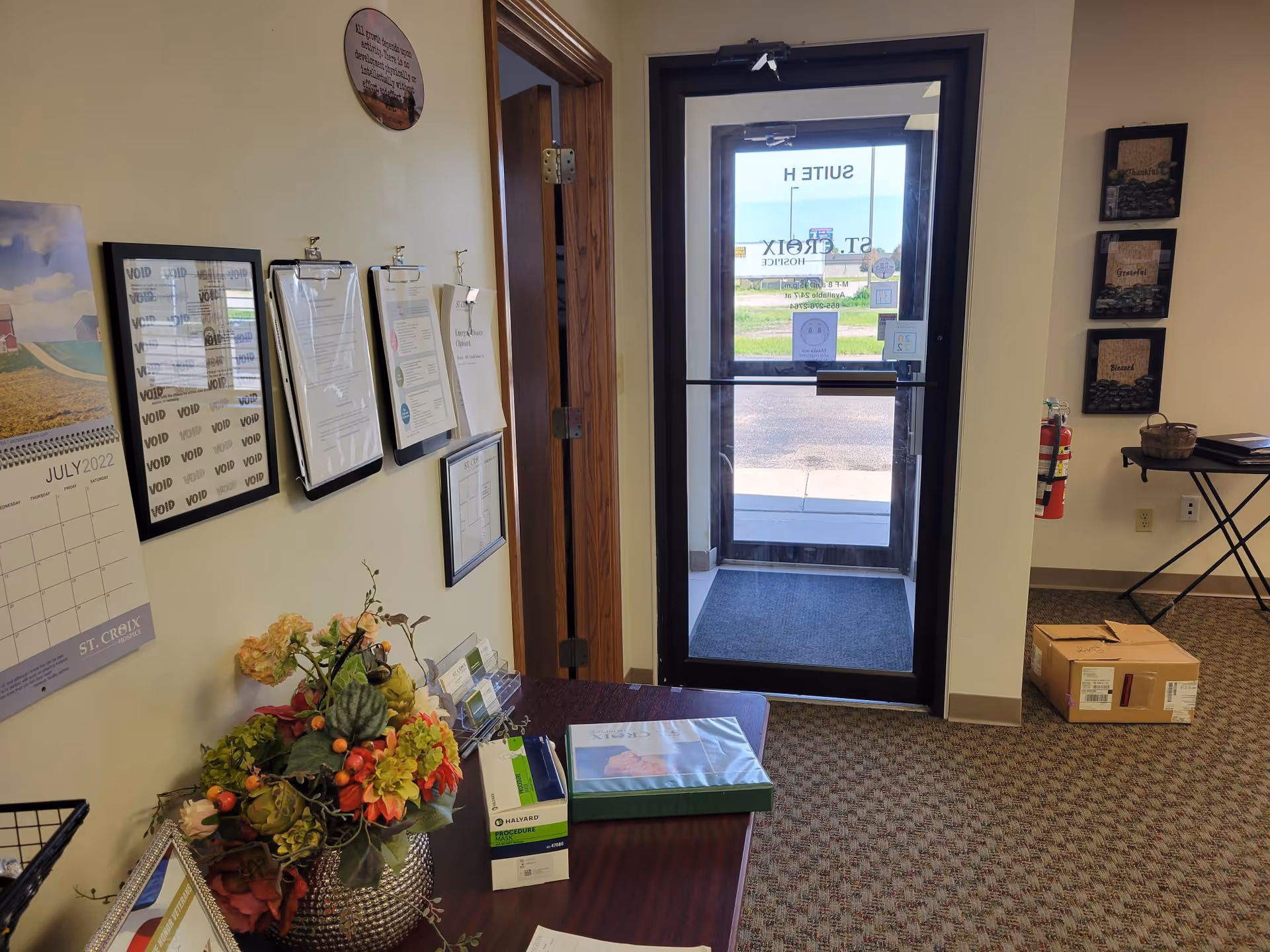 Indoor reception area with glass entry door, wall displays, desk, flowers, and a cardboard box.