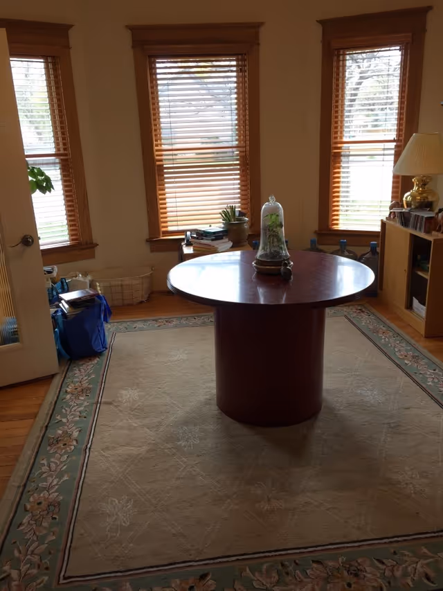 A round wooden table sits in a cozy living room with large windows and a rug.