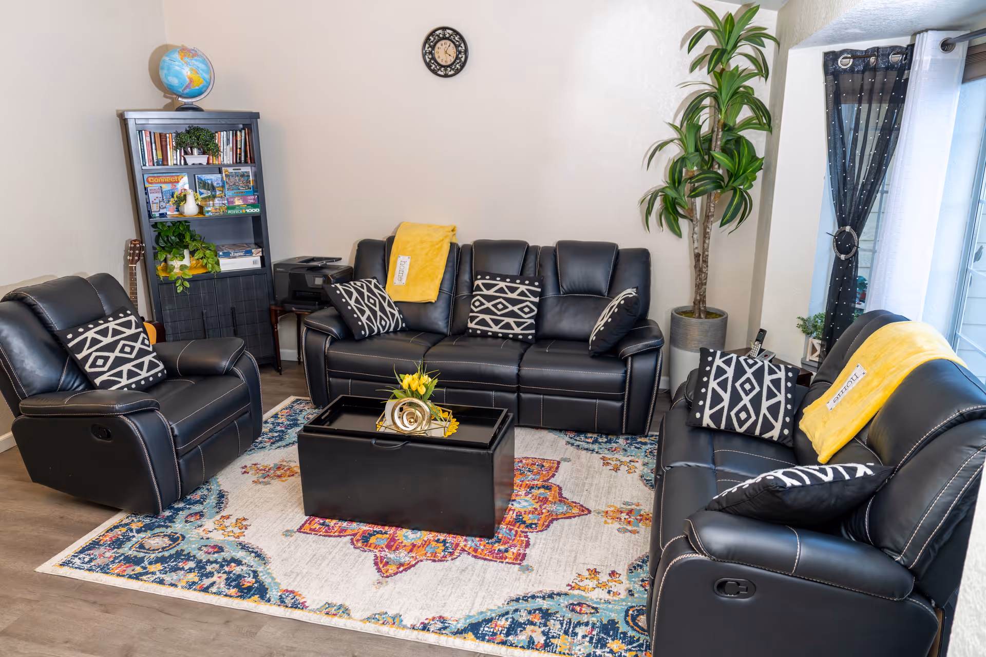 Cozy living room featuring black leather couches, pillows, a rug, plants, and a bookshelf.