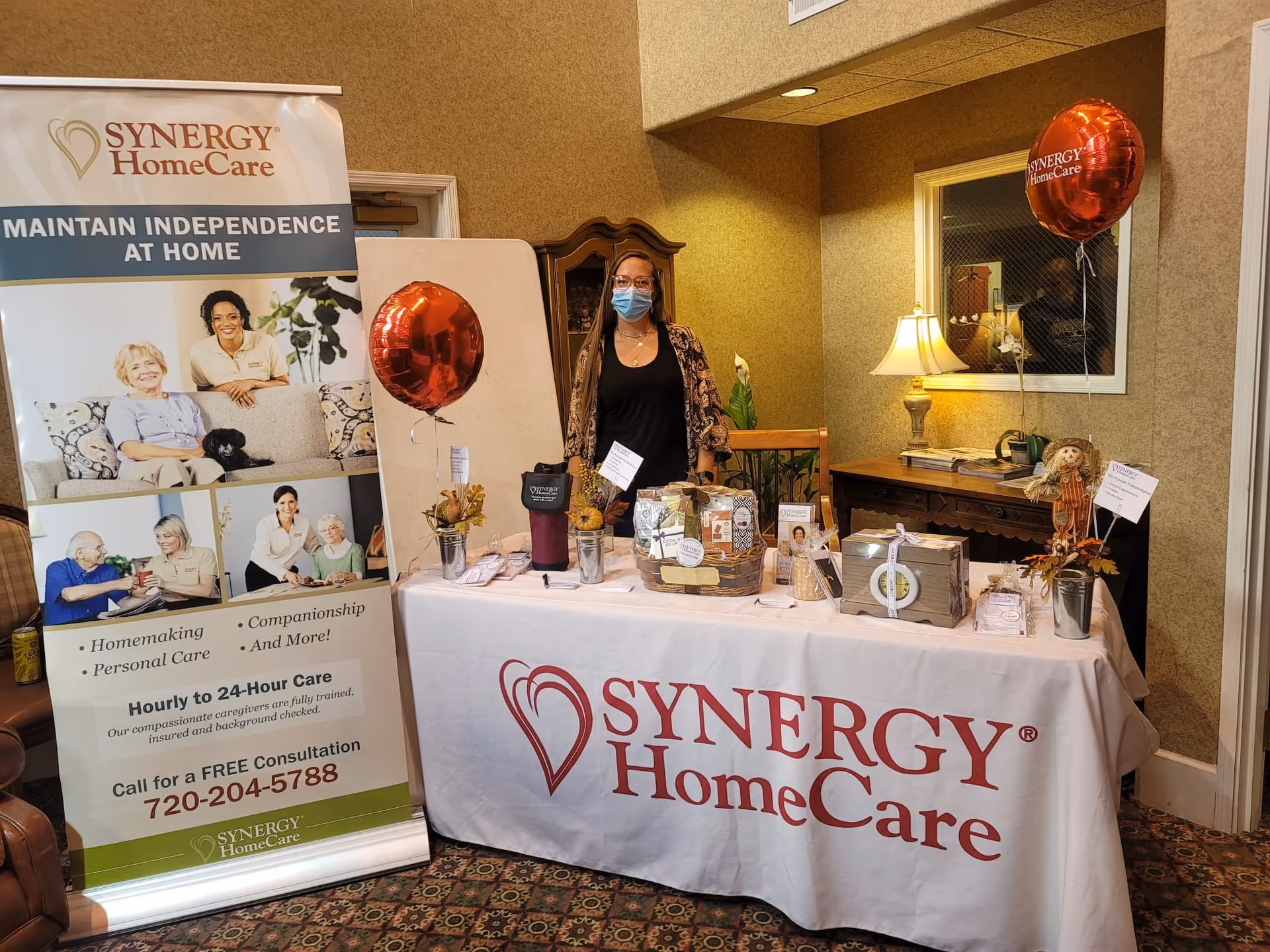 Person wearing mask stands behind a SYNERGY HomeCare table and banner in interior lobby.