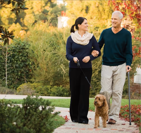 Two seniors walk a small dog outdoors on a sunny autumn day.