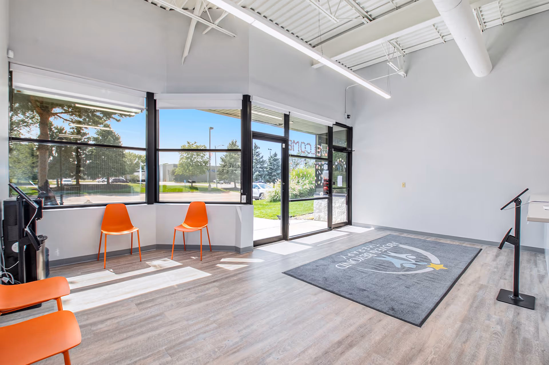 Bright interior lobby with large windows, orange chairs, and a logo mat by glass doors