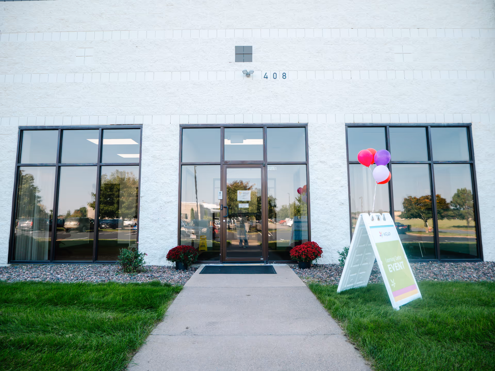 White brick building facade with large glass doors, balloons, and an outdoor signboard.