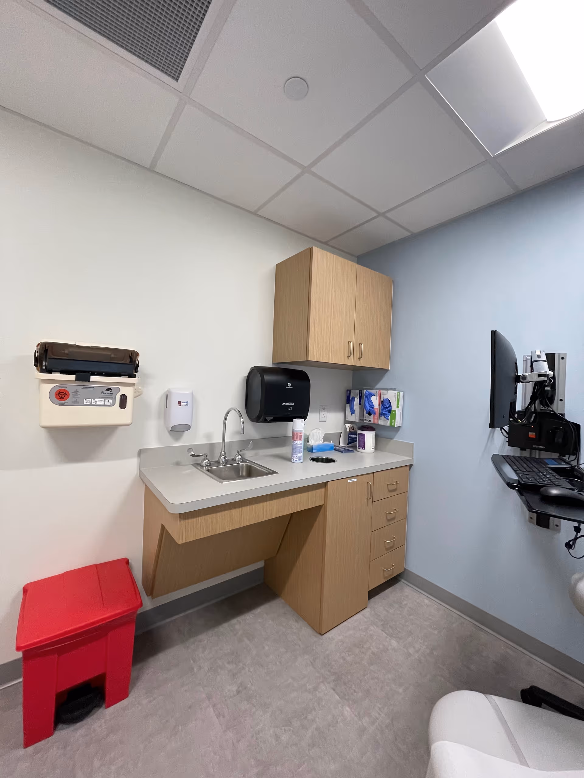 A compact clinical kitchenette with sink, counter, cabinets, and wall-mounted monitor.