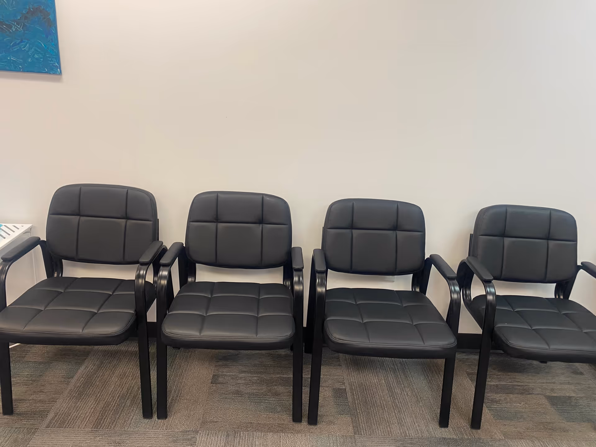 Interior waiting area with four black cushioned chairs lined up along a wall.