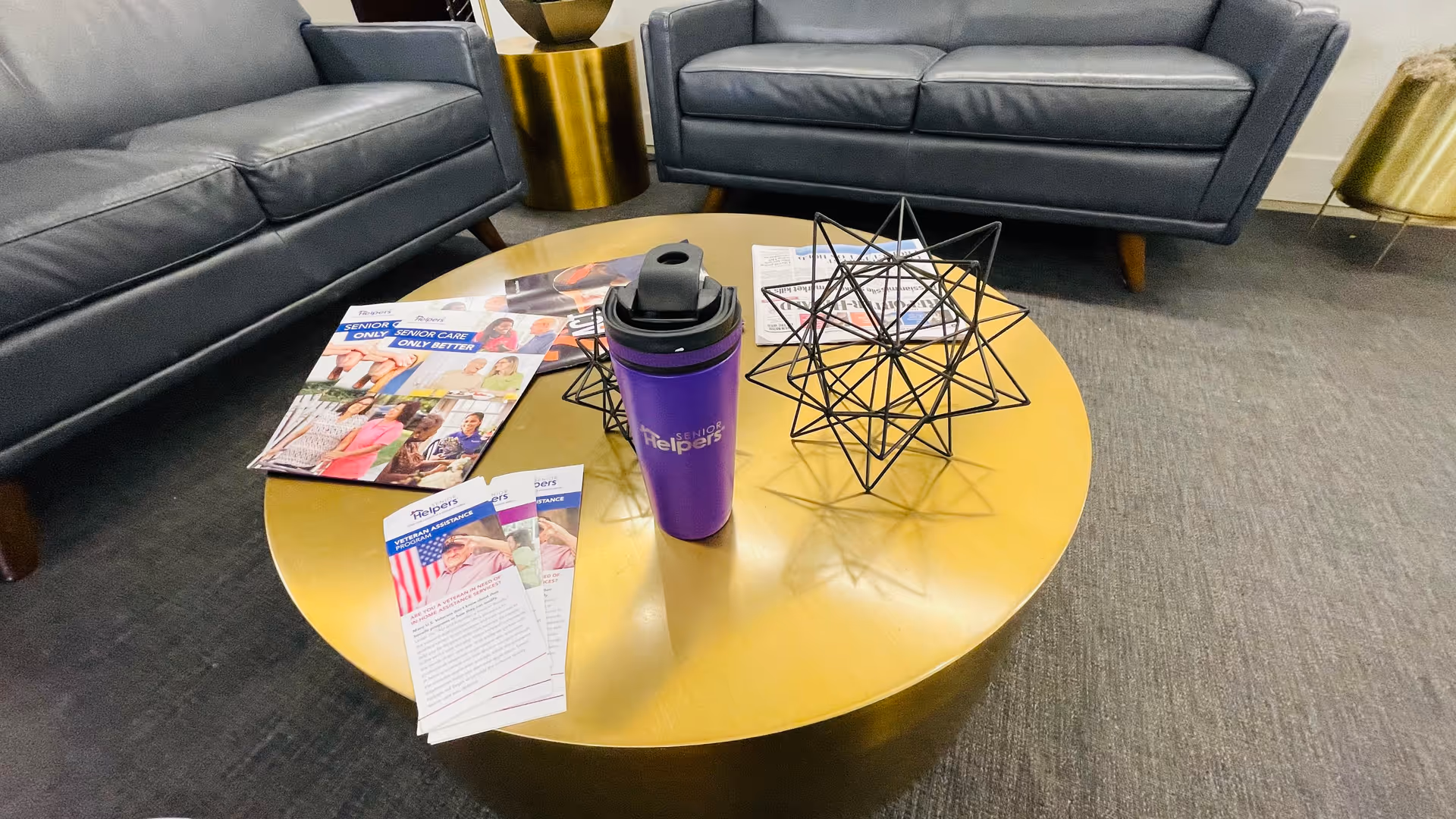A circular wood coffee table with brochures, a purple tumbler, geometric wire sculpture, and two sofas.