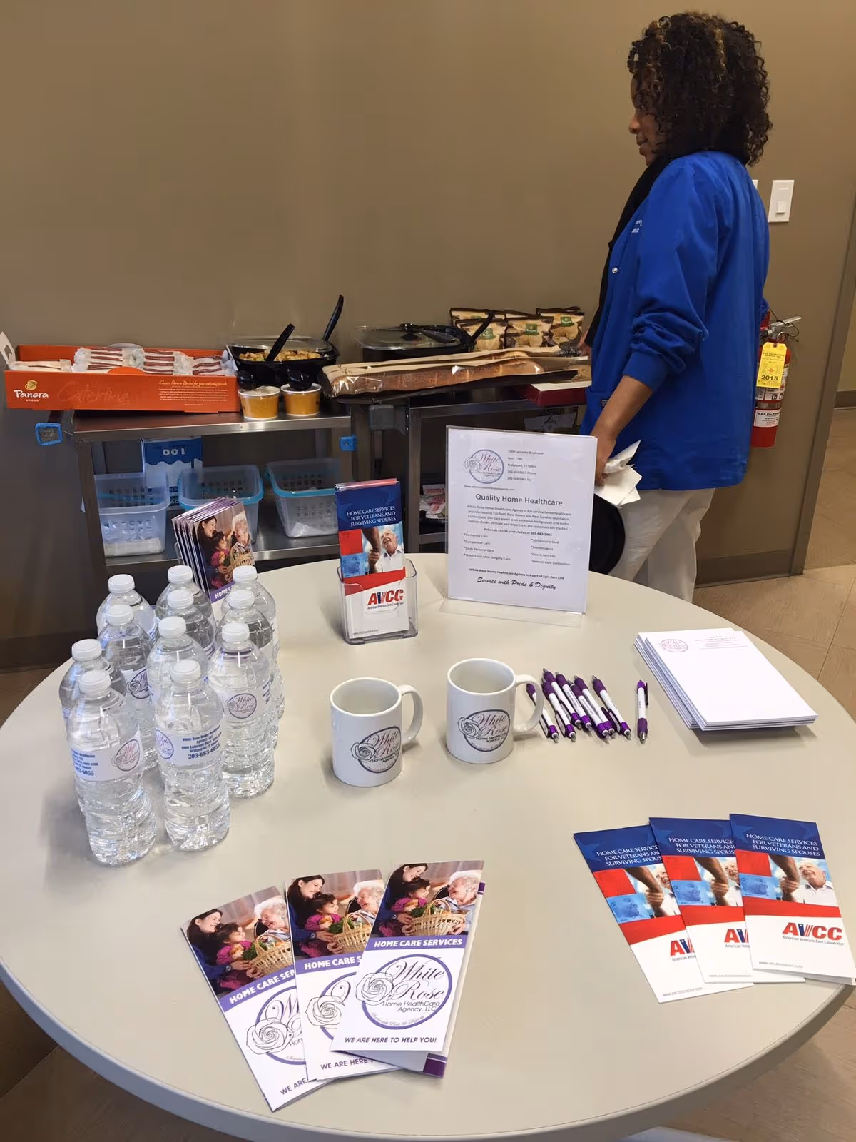 Promotional table with water bottles, brochures, and pens inside a healthcare facility, staff nearby.
