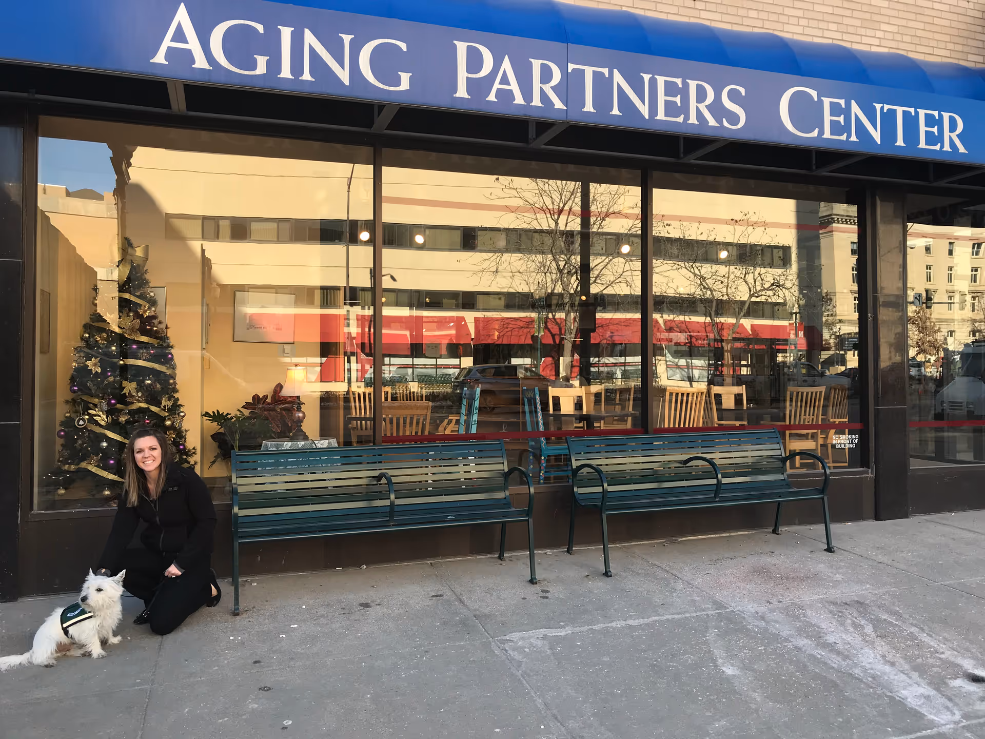 A woman kneels beside a white dog outside a storefront with benches.