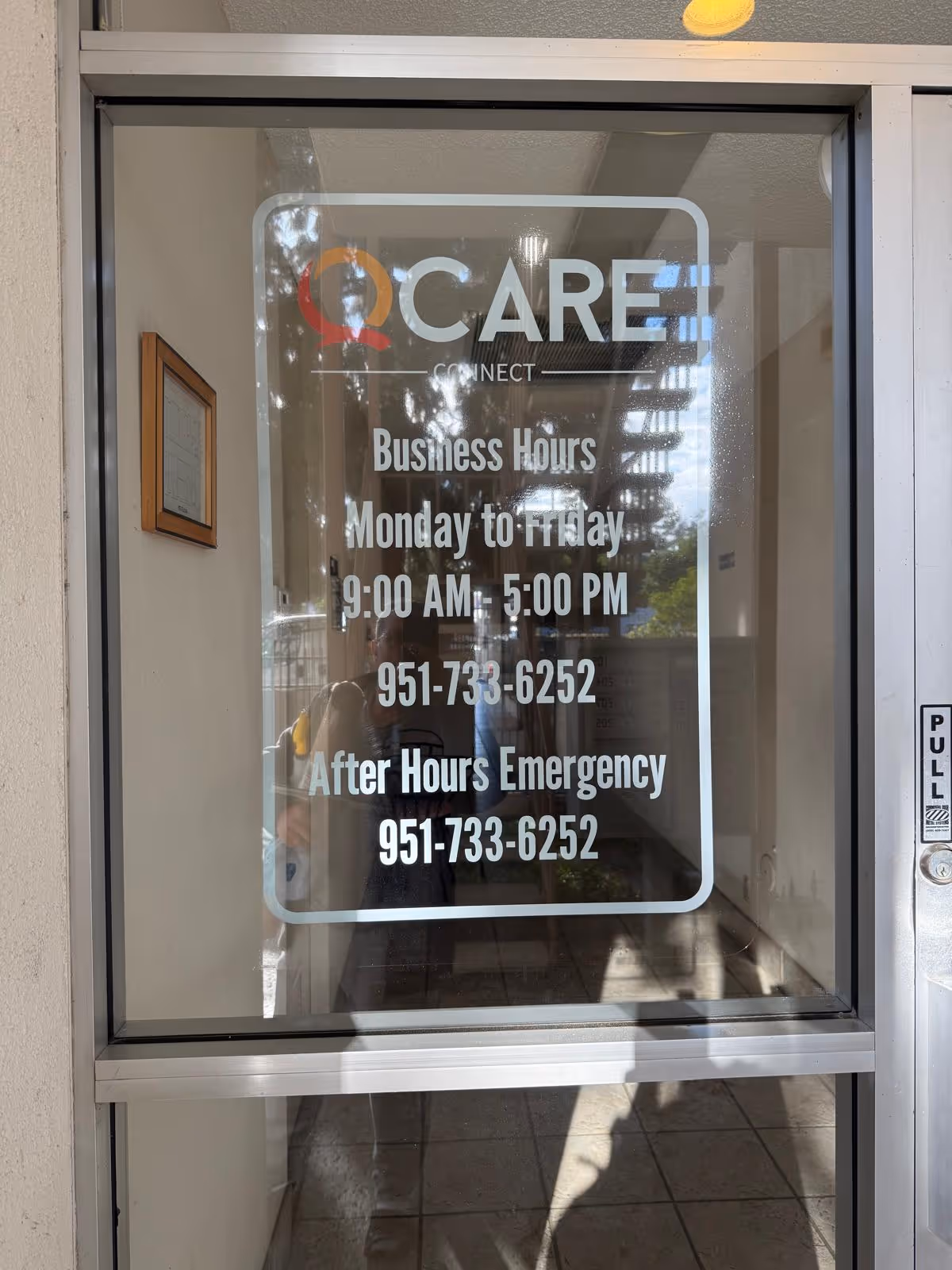 Glass door displays QCare Connect sign with business hours and emergency phone number.