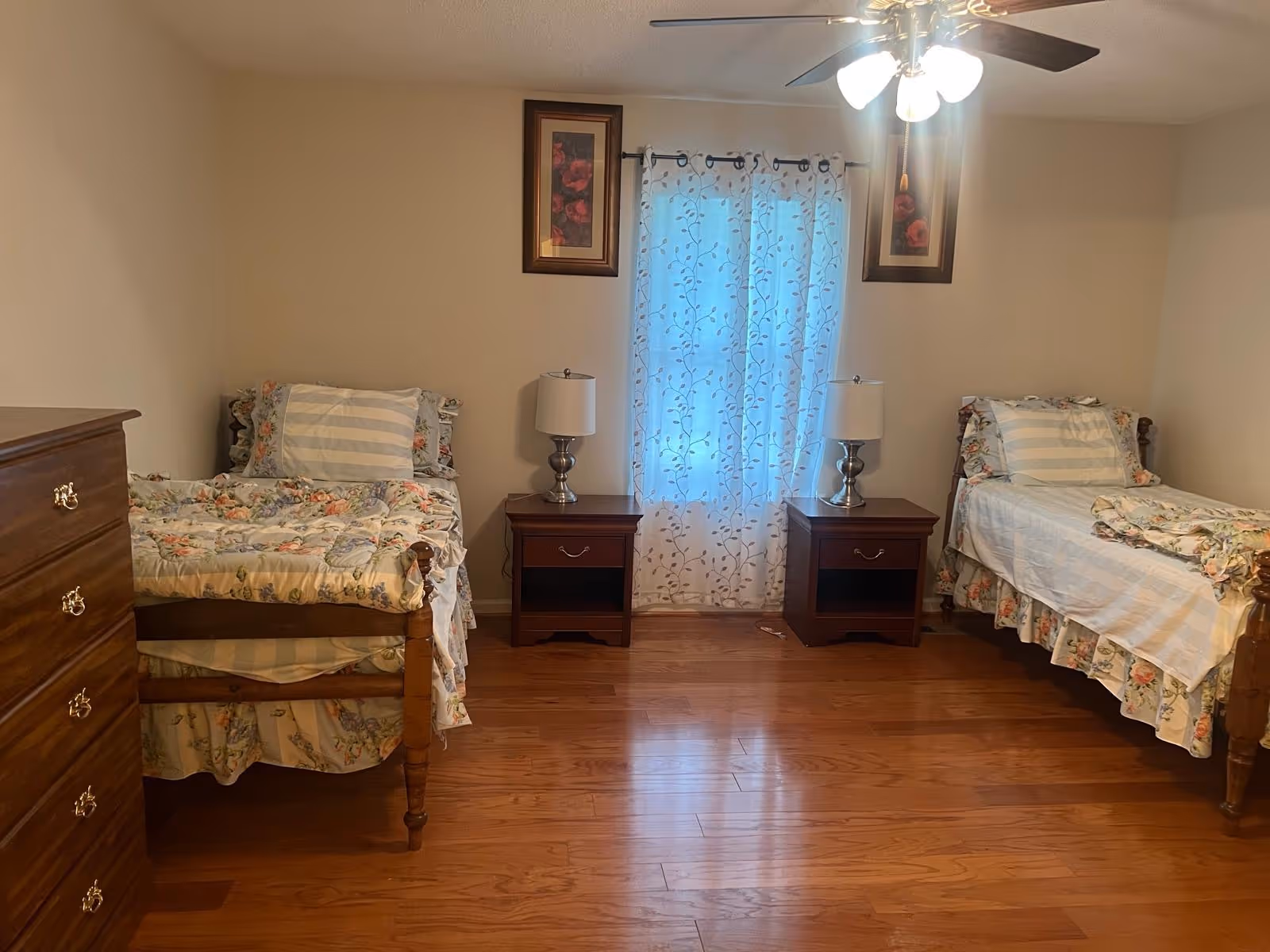 Two single beds with floral quilts, nightstands, dresser, and window with patterned curtain in a bedroom.