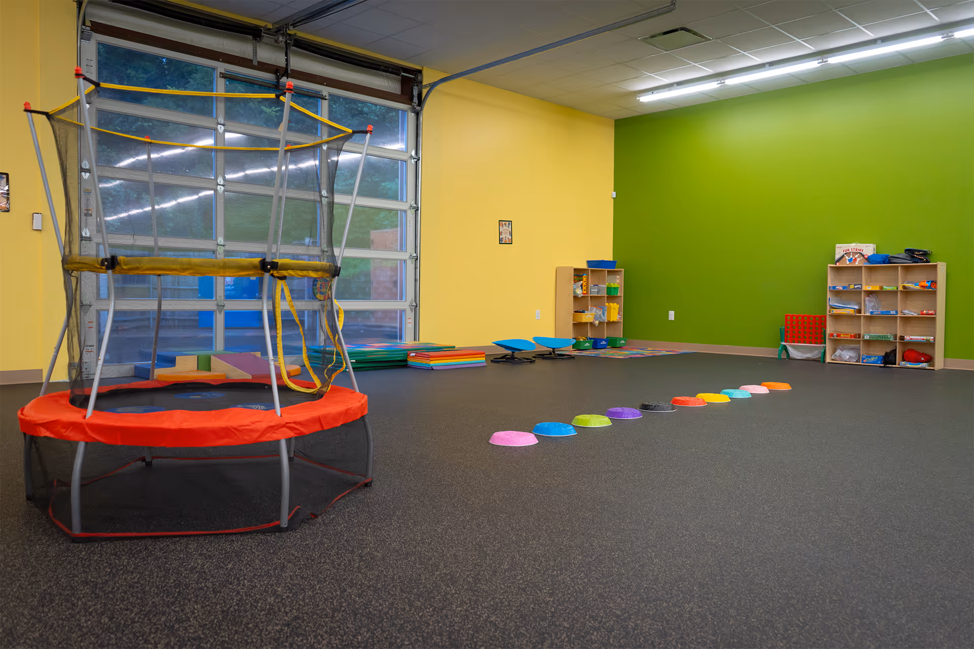 Bright therapy room with a trampoline, stepping stones, and toy shelves.