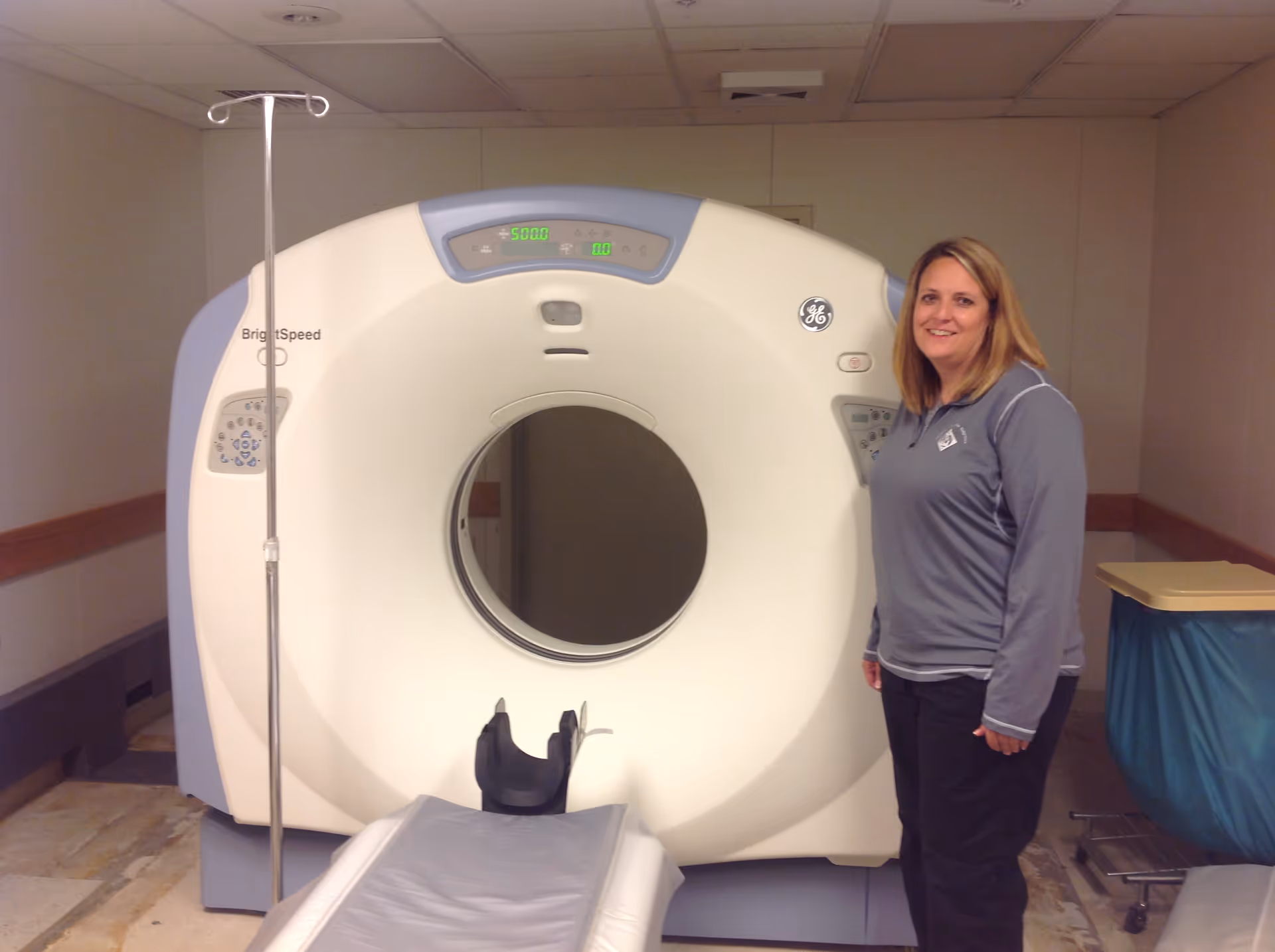 A woman stands beside a large CT scanner in a hospital interior.
