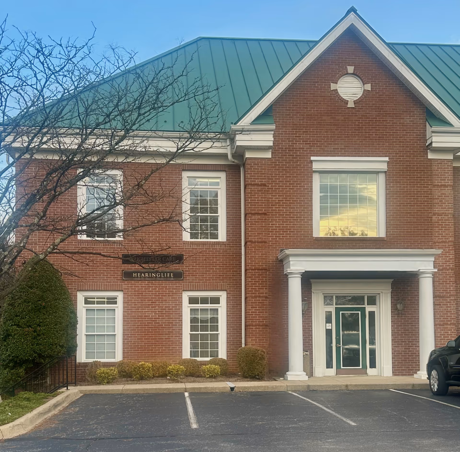 Brick building front with a green metal roof, white columns, and trimmed landscaping.