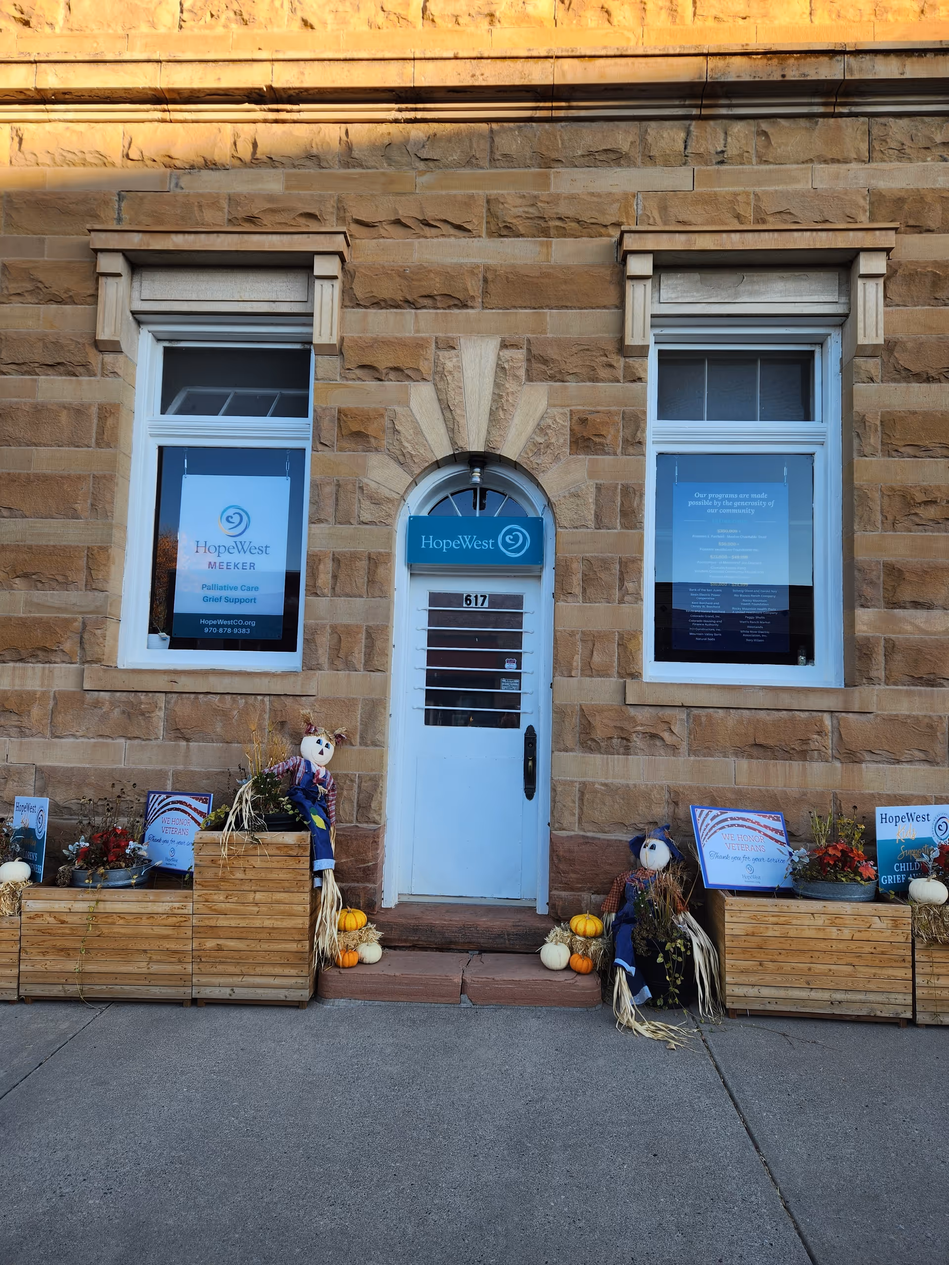 Stone building facade, central white door, blue HopeWest sign, pumpkins and planters outside.