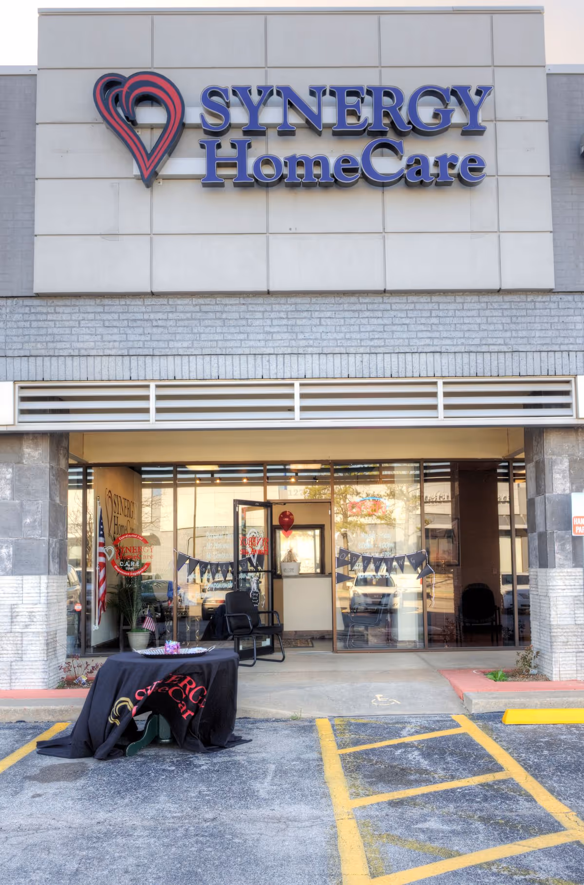 Facade of SYNERGY HomeCare facility with entrance, glass doors, and outdoor event setup.