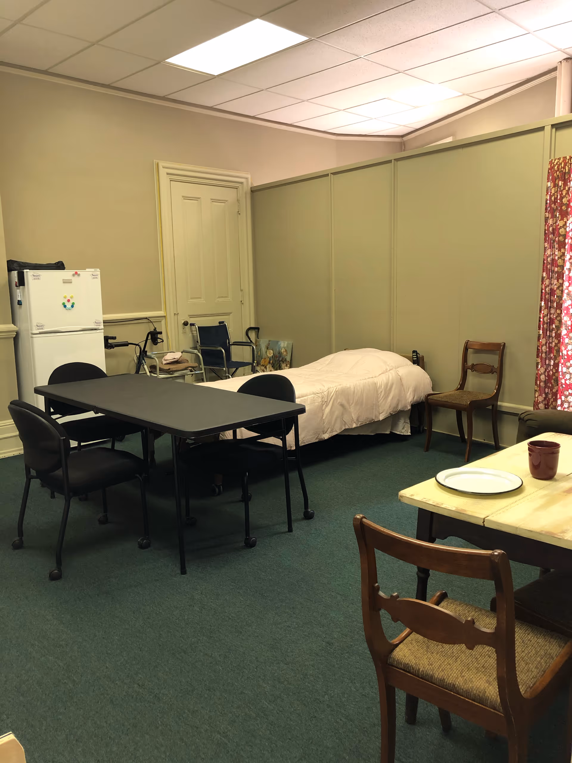 A small, sparsely furnished senior living space with a bed, two tables, chairs, and a fridge.