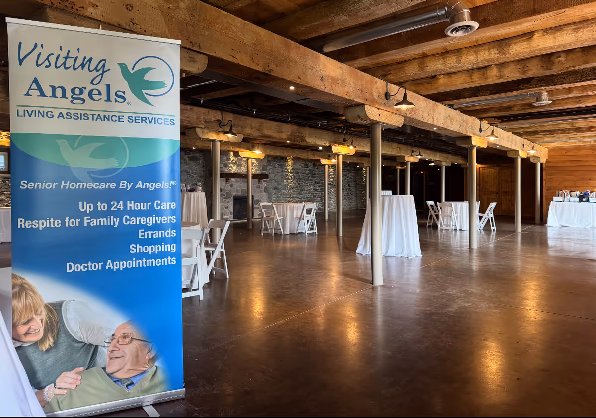 Spacious indoor event hall with wooden beams, round tables, and a large Visiting Angels banner.