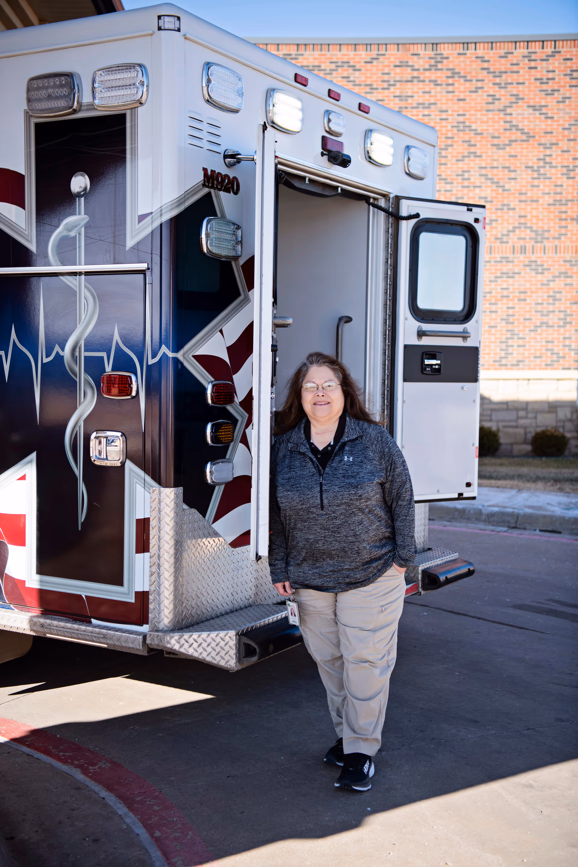 Woman stands beside an open ambulance outside a brick building.