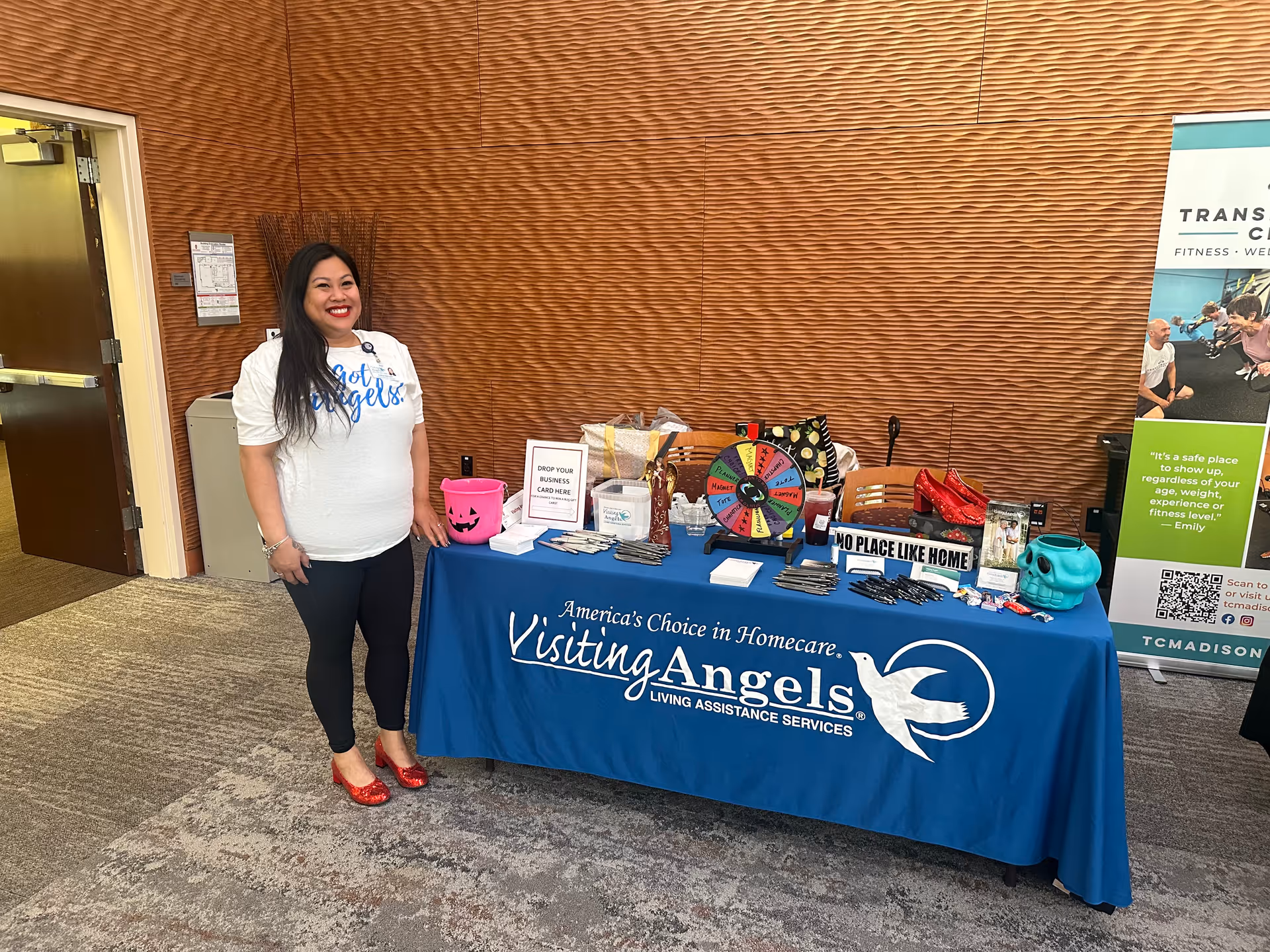 A woman stands beside a blue Visiting Angels table in a facility lobby.