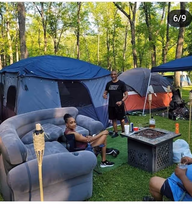Outdoor campsite with tents, seating, and a fire pit in a wooded park