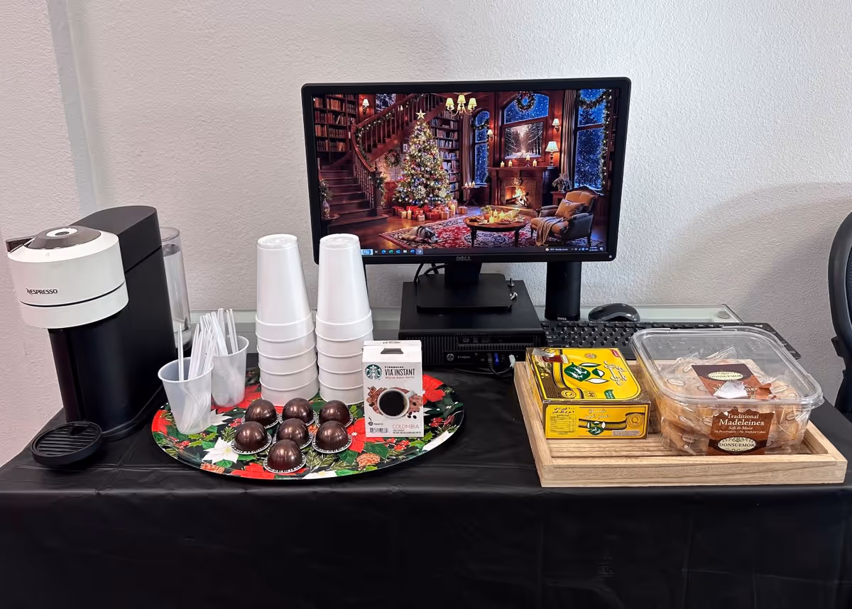 Snack station with coffee machine, cups, chocolates, and a computer monitor displaying a Christmas scene.
