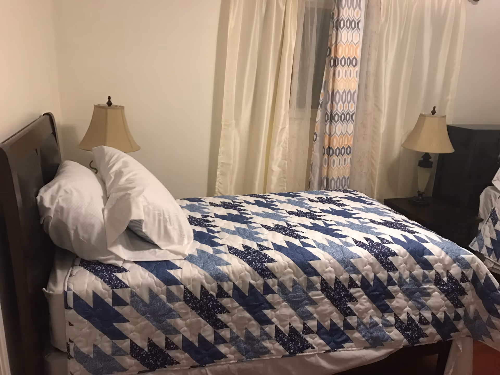 A cozy bedroom with a quilted blue-and-white bed, two lamps, and curtains.