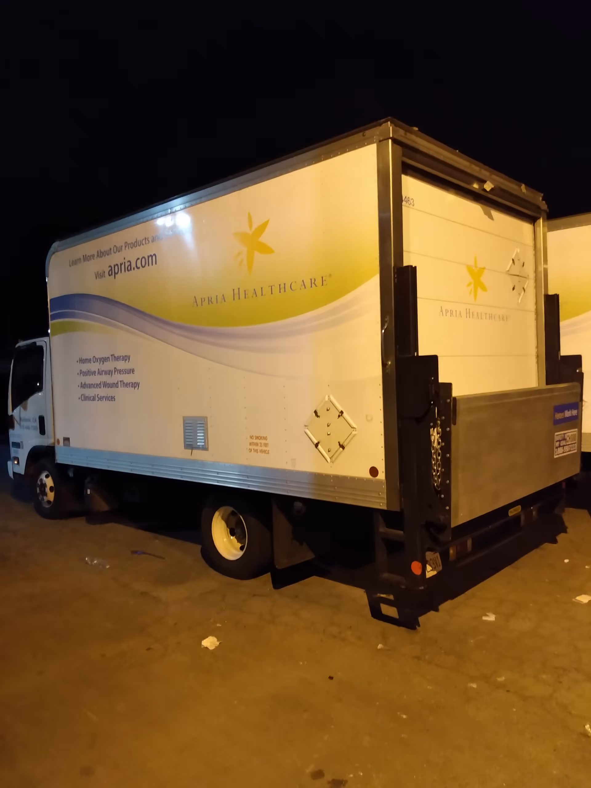 A white Apria Healthcare delivery truck trailer is parked at night.