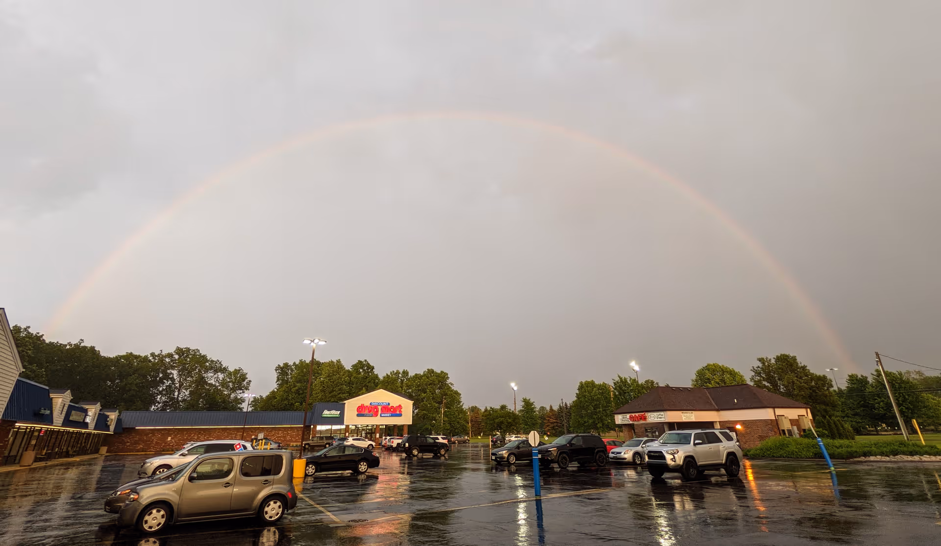 A Discount Drug Mart storefront in a parking lot after rain with a large rainbow overhead.