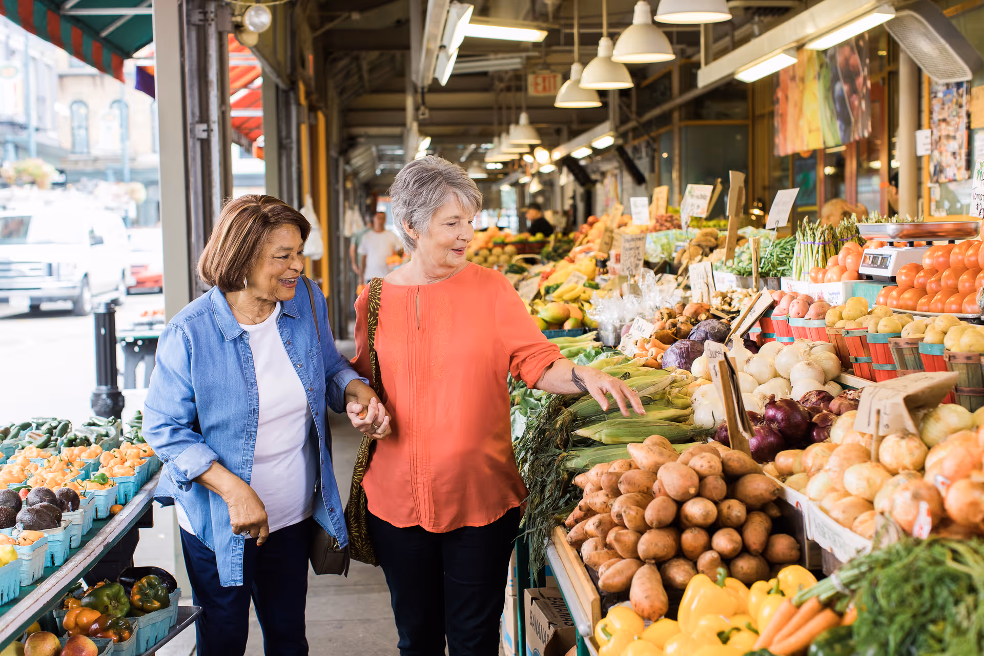 Two seniors shop at an outdoor fruit and vegetable market, smiling and pointing at produce.