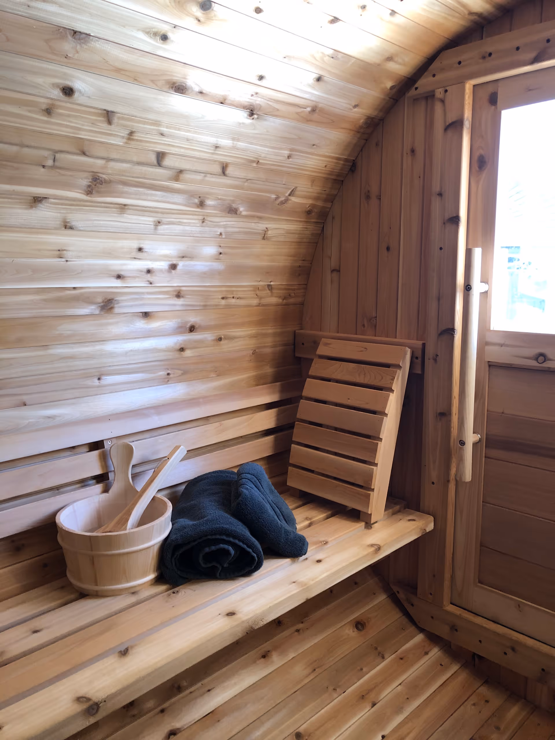 Wood-paneled sauna interior with benches, a bucket and ladle, and a folded wooden rack.