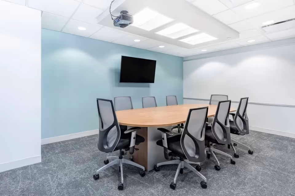 A modern interior conference room with an oval wooden table, chairs, and a wall mounted TV.