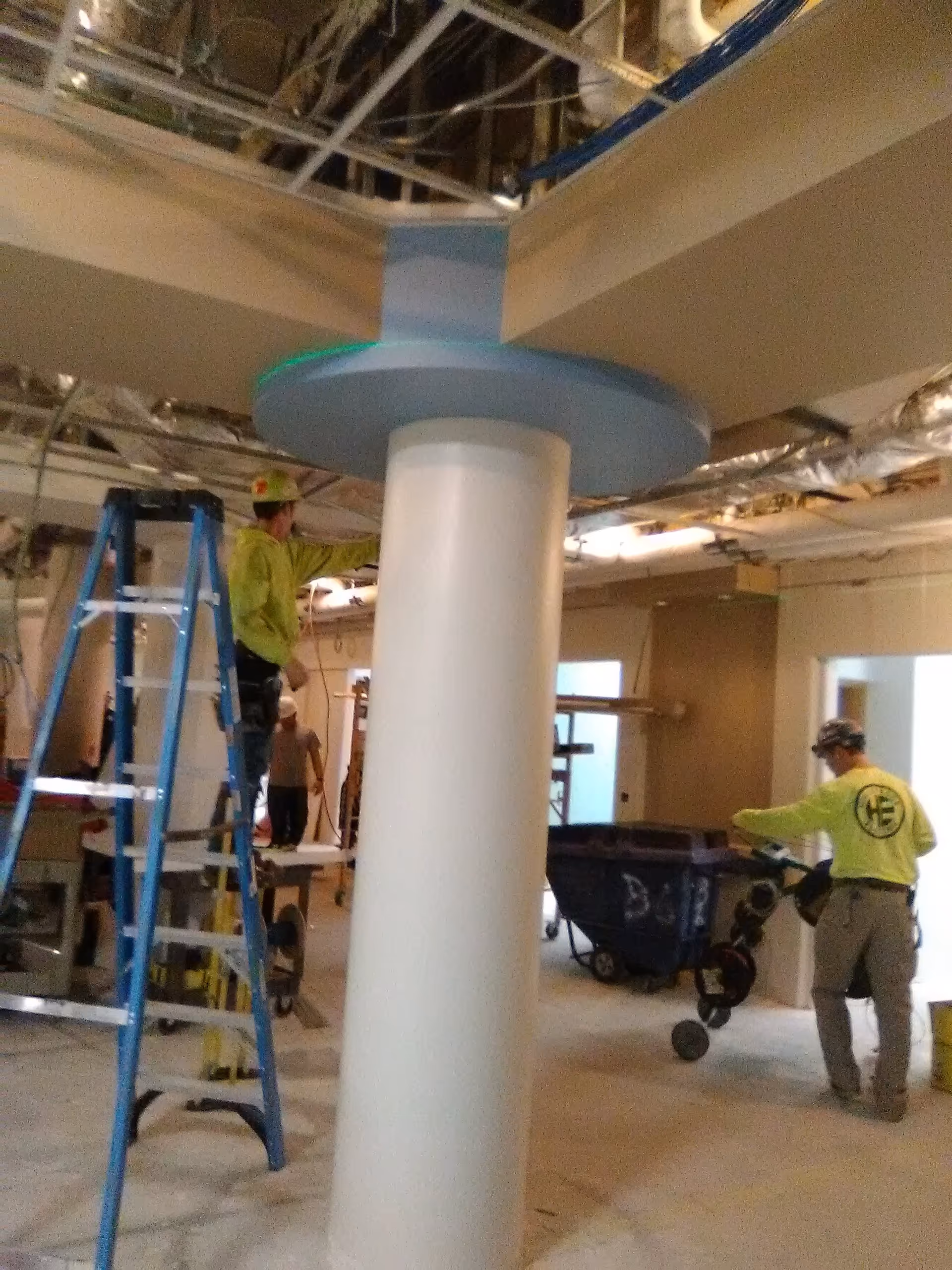 Construction workers in neon shirts renovate an indoor hospital space around a central column.