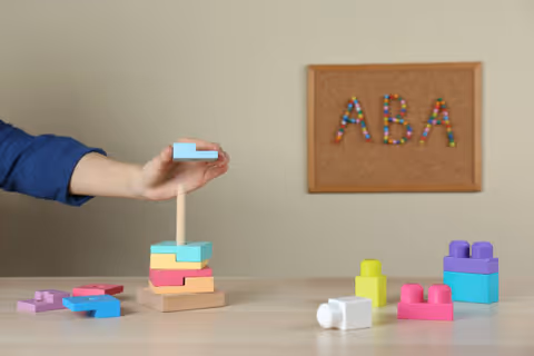 A blue-sleeved arm stacks pastel blocks on a table in a therapy room.