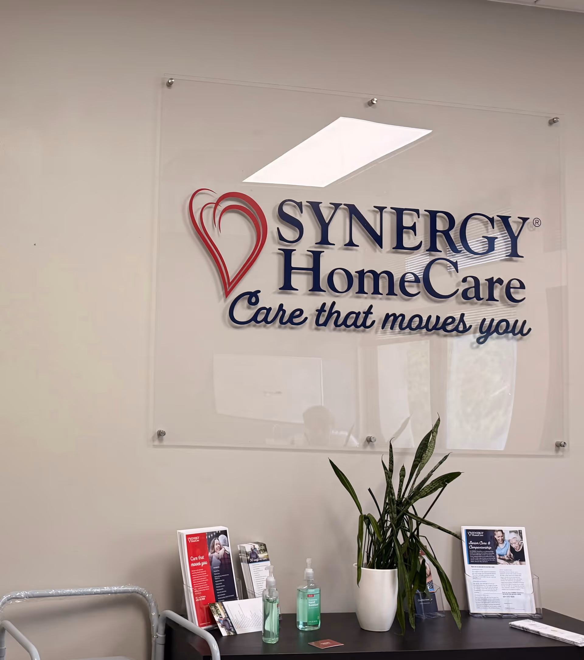 Sign reading SYNERGY HomeCare on a wall with a desk, plant, and brochures.