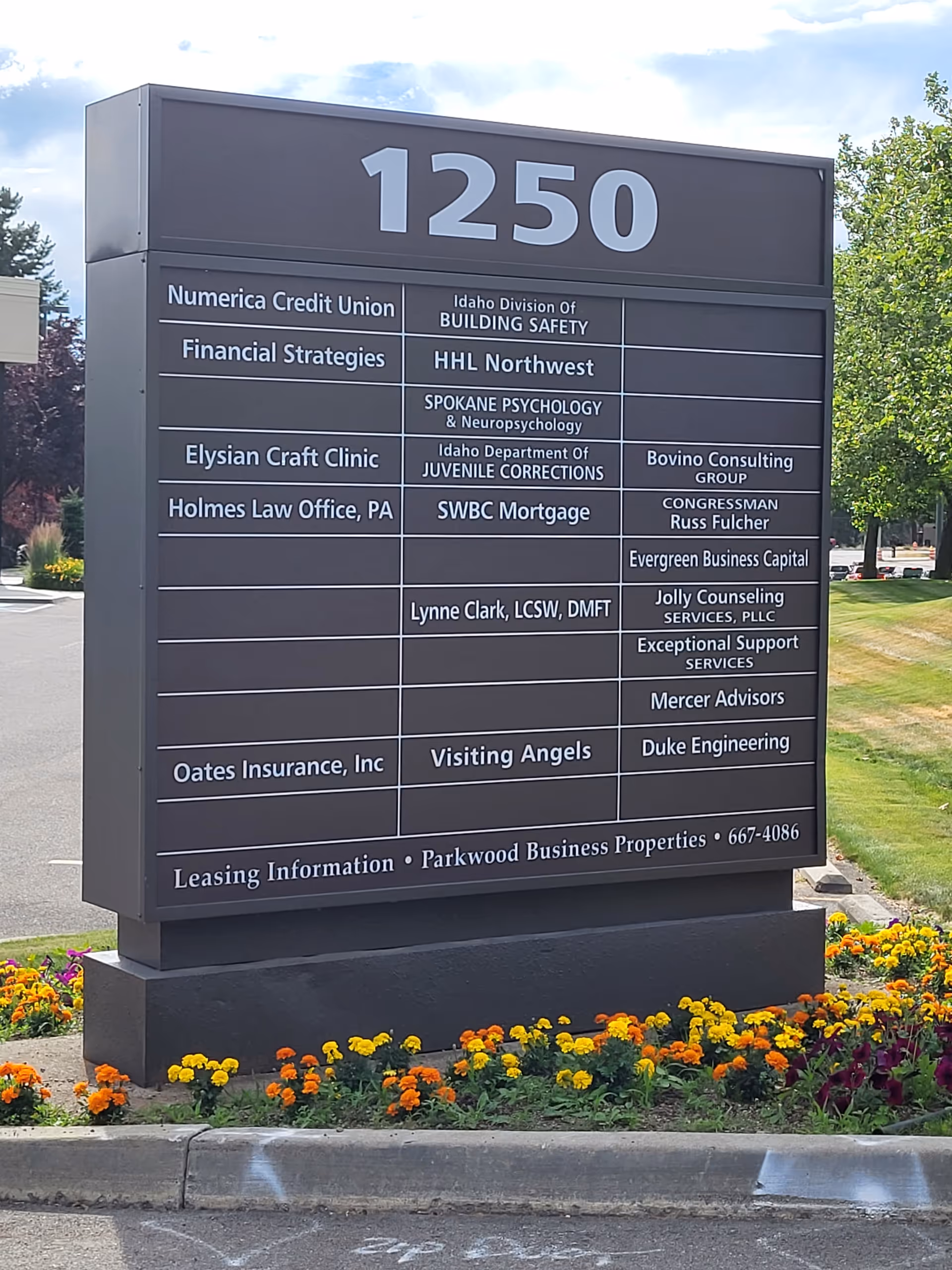 Tall outdoor directory sign displaying business names with flowers at its base