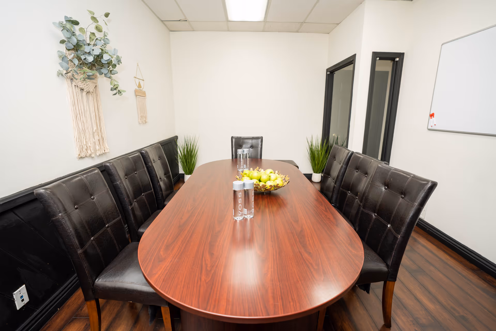 Oval wooden dining table with black upholstered chairs, fruit bowl, water bottles, and plants.