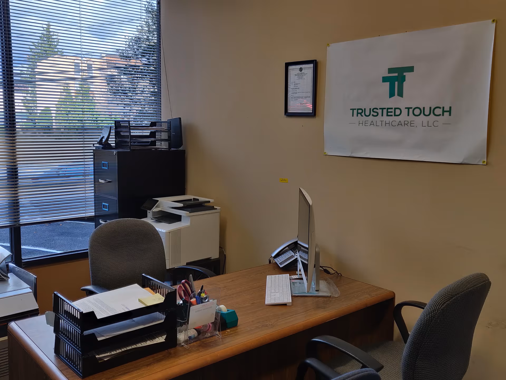 Office workspace with computer, printer, filing cabinets, and a Trusted Touch Healthcare banner on wall.