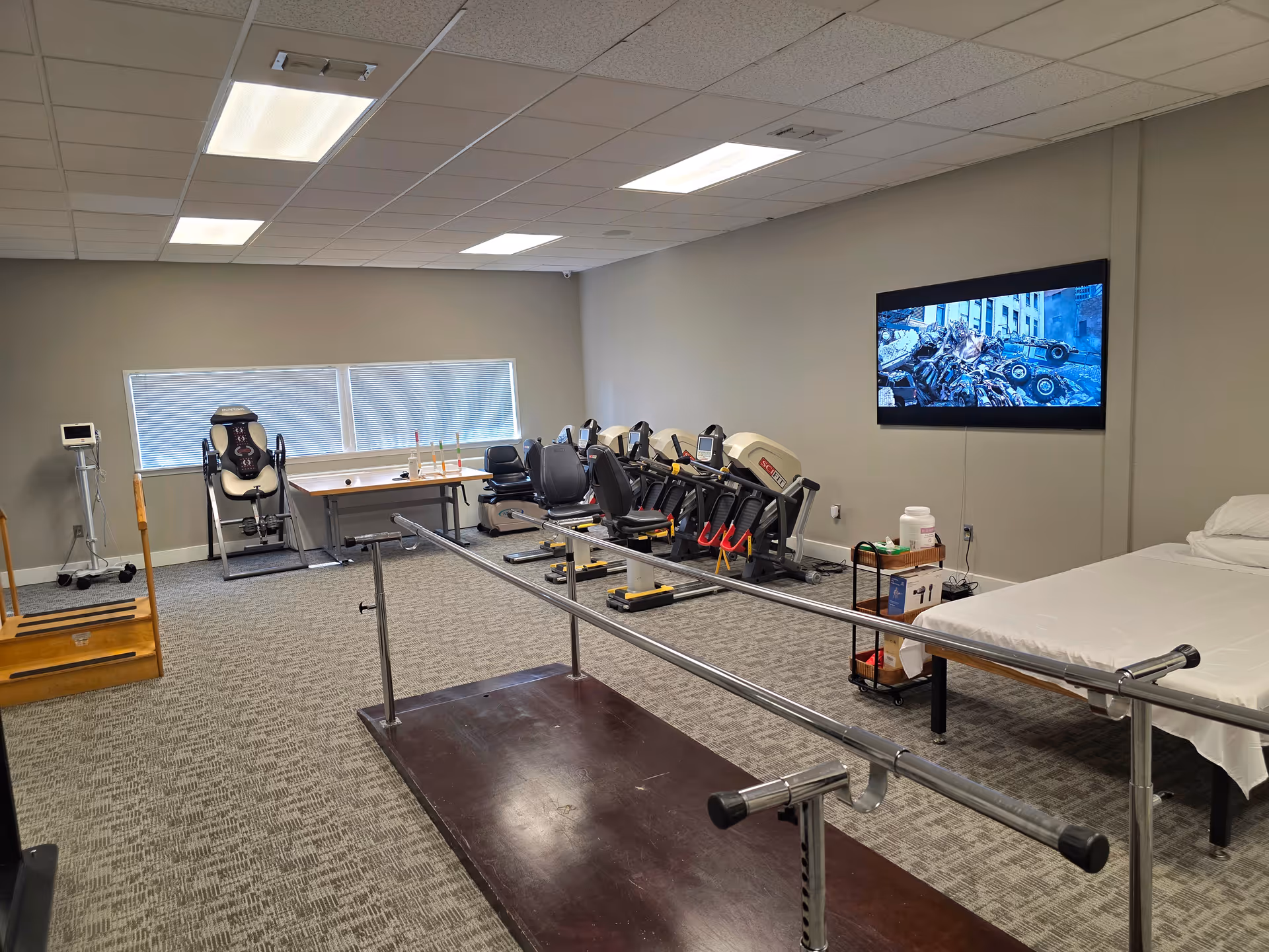 Medical therapy room with parallel bars, exercise machines, a bed, and wall-mounted TV.