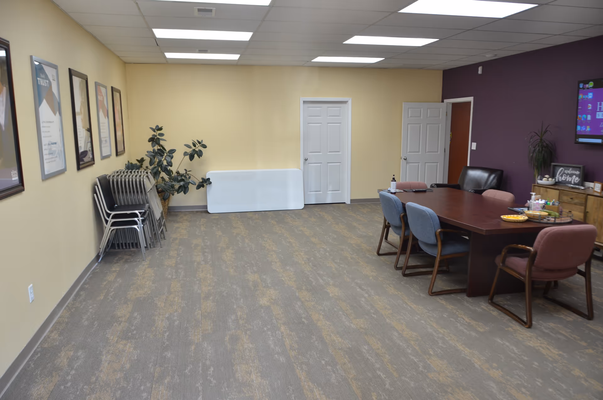 Interior senior living facility common area with table, chairs, posters, and a wall screen.