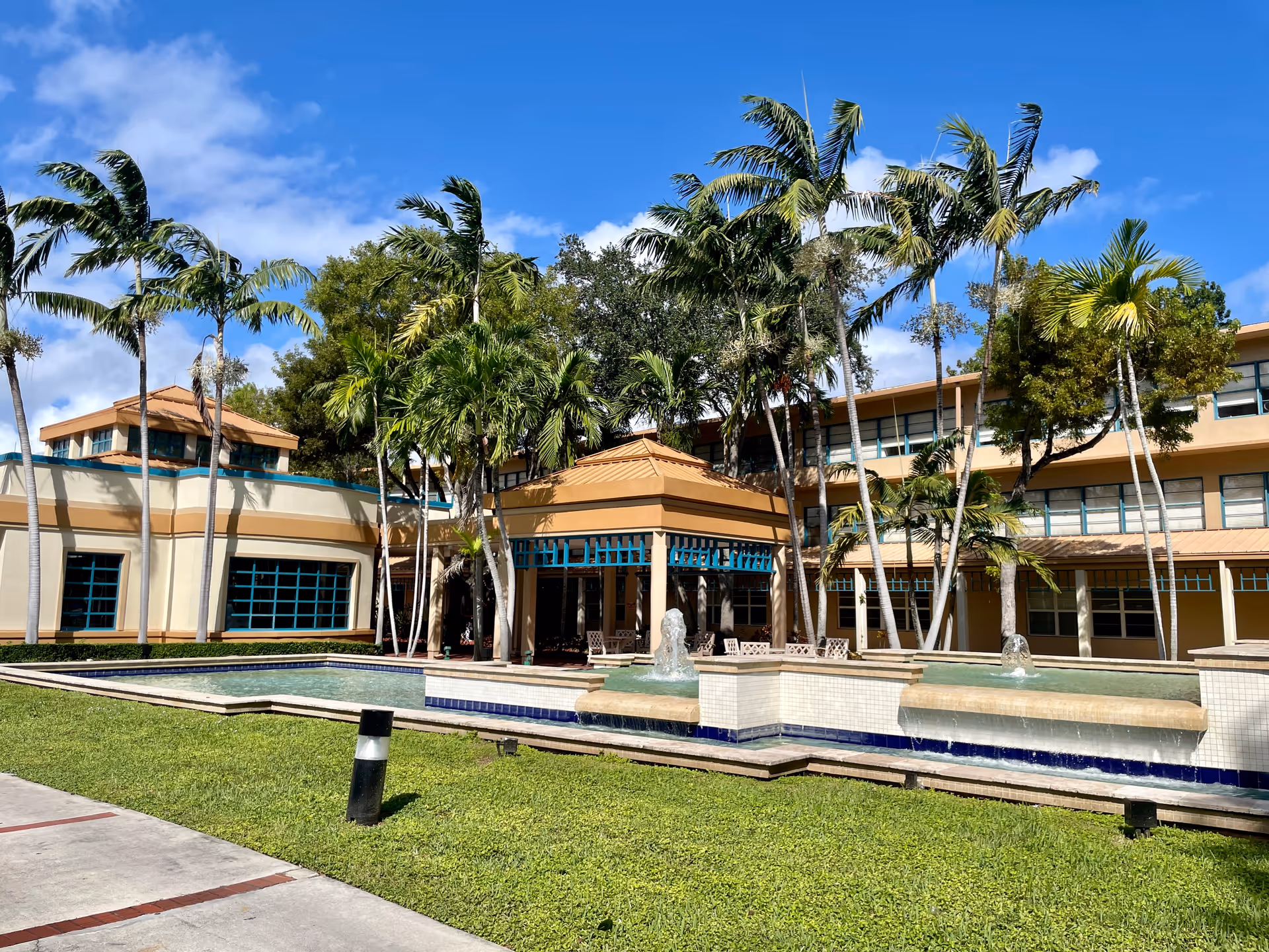 Exterior view of a beige senior facility with palm trees, courtyard fountain, and seating.