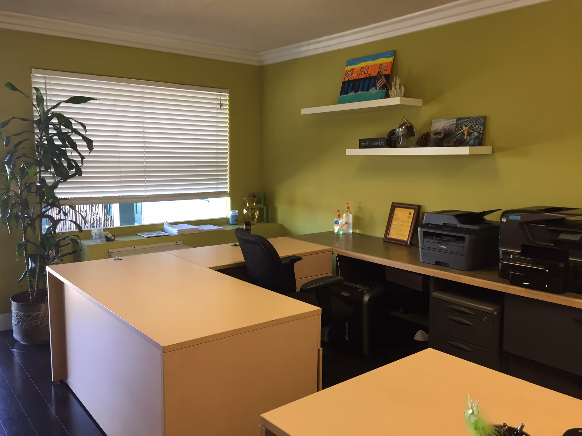 An orderly office space with L-shaped desks, a printer, and shelves along green walls.